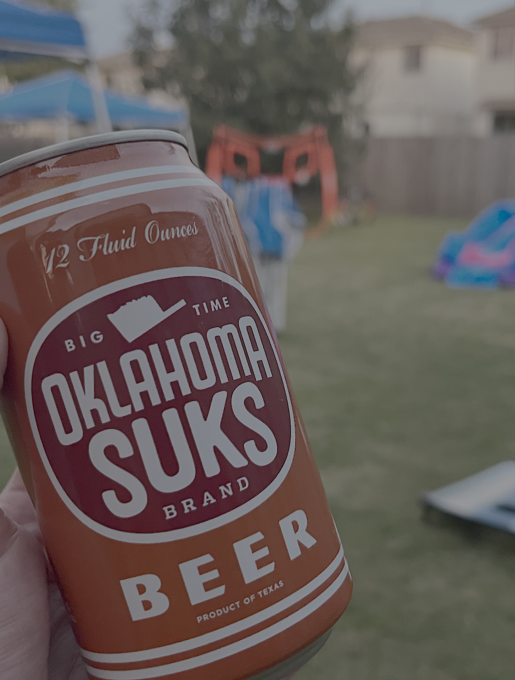A hand holds a can of beer labeled OKLAHOMA SUKS BRAND BEER with additional text 12 Fluid Ounces, BIG TIME, and PRODUCT OF TEXAS. The background shows a grassy backyard with playground equipment and houses, suggesting a casual, playful, and lighthearted outdoor gathering.